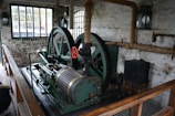 An old industrial machine with large green metal gears and a red handle is situated in a brick-walled room. There are tall windows with black frames, allowing natural light to enter. The walls have exposed piping wrapped in insulation, and a wooden railing borders the machinery.
