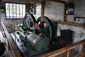 An old industrial machine with large green metal gears and a red handle is situated in a brick-walled room. There are tall windows with black frames, allowing natural light to enter. The walls have exposed piping wrapped in insulation, and a wooden railing borders the machinery.