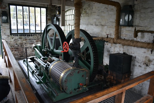 An old industrial machine with large green metal gears and a red handle is situated in a brick-walled room. There are tall windows with black frames, allowing natural light to enter. The walls have exposed piping wrapped in insulation, and a wooden railing borders the machinery.