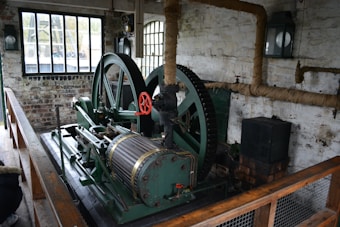 An old industrial machine with large green metal gears and a red handle is situated in a brick-walled room. There are tall windows with black frames, allowing natural light to enter. The walls have exposed piping wrapped in insulation, and a wooden railing borders the machinery.
