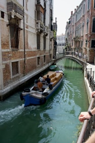 A narrow canal in an urban setting featuring a small cargo boat carrying several boxes. The canal is flanked by tall, historic buildings with weathered facades. A small arched bridge crosses the canal in the distance, and there are people walking on it. The water is a greenish hue, and the scene suggests transportation in a historic city.