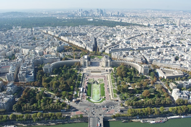 An expansive aerial view of a large city, characterized by dense urban architecture. A prominent green space with neatly arranged gardens and wide pathways is centered in the foreground. The urban landscape extends into the distance, revealing a mix of historical and modern buildings, with a cluster of tall skyscrapers at the horizon.