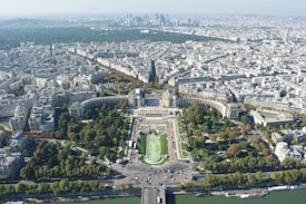 An expansive aerial view of a large city, characterized by dense urban architecture. A prominent green space with neatly arranged gardens and wide pathways is centered in the foreground. The urban landscape extends into the distance, revealing a mix of historical and modern buildings, with a cluster of tall skyscrapers at the horizon.