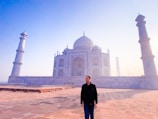 A person stands in front of the Taj Mahal, an iconic white marble mausoleum with a large dome and four surrounding minarets. The sky is clear with a light gradient of blues and the early morning sun casts a soft glow over the structure and the stone courtyard.