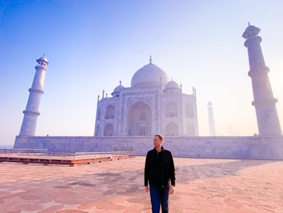 A person stands in front of the Taj Mahal, an iconic white marble mausoleum with a large dome and four surrounding minarets. The sky is clear with a light gradient of blues and the early morning sun casts a soft glow over the structure and the stone courtyard.