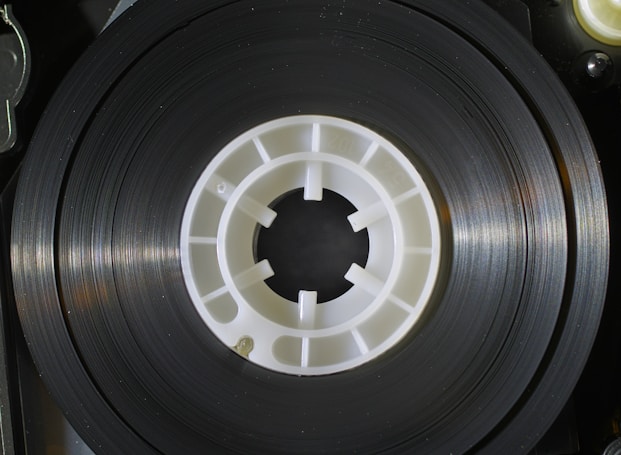 A close-up image of a reel of magnetic tape, featuring a black circular tape with a white plastic hub at the center. The surface of the tape is shiny and reflects light, while the hub has evenly spaced spokes extending outward from the center.
