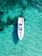 Aerial view of a white yacht floating in clear turquoise water. The surrounding water reveals underwater rocks and formations, creating a pattern beneath the surface. A person is visible sunbathing on the deck of the yacht.