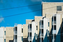 A modern apartment building with a series of vertical and horizontal lines, featuring multiple balconies and beige-colored exterior walls. The building is set against a bright blue sky with a few clouds, and there are electrical wires crossing the scene.