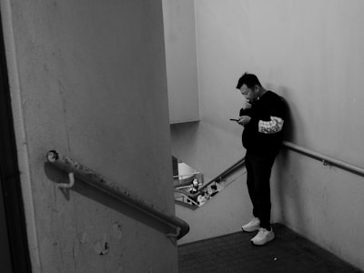 A person is standing at the corner of a staircase, looking at a smartphone. The setting is indoors with plain walls and metal railings. The atmosphere appears quiet and individual-focused.