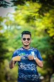 A man is standing outdoors with sunglasses on, wearing a sports jersey. The jersey features a flag patch and a crest on the front. The background is a blurred scene of green foliage, giving a natural and serene setting.