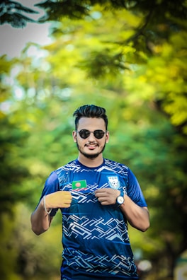A man is standing outdoors with sunglasses on, wearing a sports jersey. The jersey features a flag patch and a crest on the front. The background is a blurred scene of green foliage, giving a natural and serene setting.
