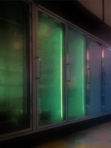 A row of industrial-style glass doors displaying a greenish-blue glow, suggesting refrigeration units in a dimly lit environment. The surfaces of the glass doors have a frosty or misted appearance, reflecting neon lights from the interior.