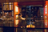 A person wearing a hat and coat is standing in a cozy bookstore, surrounded by shelves filled with books. Warm lighting casts reflections on a glass surface, creating an intimate and serene atmosphere. Framed pictures are visible on orange walls, enhancing the artistic and cultured environment.