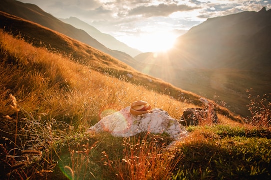 Golden light from the setting sun bathes a mountainous landscape, casting a warm glow over the grassy hills. A hat rests on a prominent rock in the foreground, adding a human touch to the natural scene. Shadows from distant peaks create a serene and contemplative atmosphere.