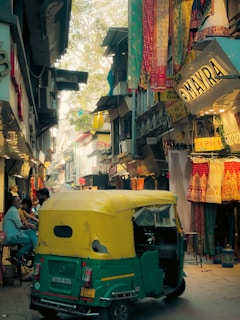 A narrow, bustling street market lined with colorful textiles and garments. An auto-rickshaw with yellow and green colors is parked in the foreground. People are sitting and chatting on the left side. Various signs and shop banners are visible, adding to the lively atmosphere. Overhead fabrics and decorations hang from the buildings, contributing to the vibrant environment.