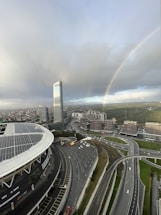 An urban landscape with a prominent high-rise building and large stadium featuring solar panels. There is a complex network of highways below, and a vibrant rainbow arcs across a cloudy sky. In the background, numerous smaller buildings are surrounded by patches of greenery.