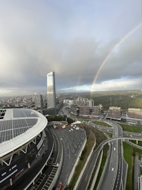An urban landscape with a prominent high-rise building and large stadium featuring solar panels. There is a complex network of highways below, and a vibrant rainbow arcs across a cloudy sky. In the background, numerous smaller buildings are surrounded by patches of greenery.