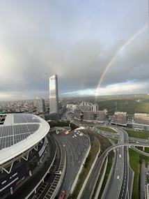 An urban landscape with a prominent high-rise building and large stadium featuring solar panels. There is a complex network of highways below, and a vibrant rainbow arcs across a cloudy sky. In the background, numerous smaller buildings are surrounded by patches of greenery.