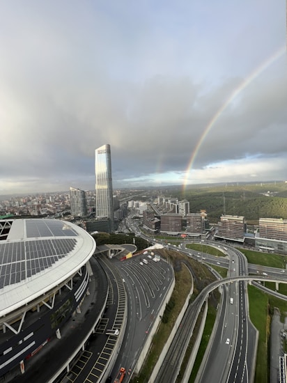 An urban landscape with a prominent high-rise building and large stadium featuring solar panels. There is a complex network of highways below, and a vibrant rainbow arcs across a cloudy sky. In the background, numerous smaller buildings are surrounded by patches of greenery.