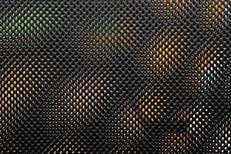 A close-up view of a mesh fabric. The pattern consists of small, evenly spaced holes creating a net-like effect. Through the mesh, hints of green and orange colors are visible, possibly indicating objects or surfaces behind it.
