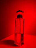 A sleek, rectangular perfume bottle with a square cap is set against a vivid red background. The transparent bottle reveals a faint liquid inside and features the text 'after five' and '5th avenue' printed vertically on its surface. The lighting creates a dramatic shadow beneath the bottle, enhancing its elegant design.