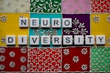 Scrabble tiles spell out 'neuro diversity' against a vibrant, ornate background composed of colorful square tiles with floral and geometric patterns.