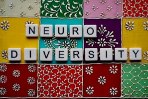 Scrabble tiles spell out 'neuro diversity' against a vibrant, ornate background composed of colorful square tiles with floral and geometric patterns.