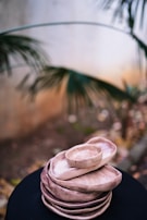 A stack of handmade ceramic plates and bowls in earthy tones, placed on a dark surface. The background features blurred foliage, giving a natural outdoor ambiance.