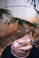A stack of handmade ceramic plates and bowls in earthy tones, placed on a dark surface. The background features blurred foliage, giving a natural outdoor ambiance.
