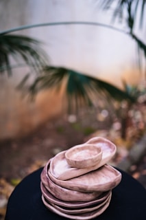 A stack of handmade ceramic plates and bowls in earthy tones, placed on a dark surface. The background features blurred foliage, giving a natural outdoor ambiance.