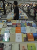 A bookstore or library setting with numerous books arranged on large tables, covering a wide range of topics and genres. People are browsing the books, creating a bustling atmosphere. The shelves in the background are filled with even more books, indicating a rich variety of reading material. Lighting is warm, enhancing the inviting ambiance.
