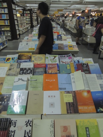 A bookstore or library setting with numerous books arranged on large tables, covering a wide range of topics and genres. People are browsing the books, creating a bustling atmosphere. The shelves in the background are filled with even more books, indicating a rich variety of reading material. Lighting is warm, enhancing the inviting ambiance.