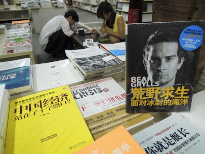 A cluttered bookstore counter with various books in Chinese and English, prominently featuring a book by Bear Grylls. Two individuals are seated in the background, engaged in reading or writing.