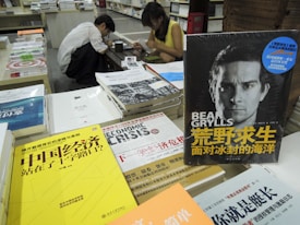 A cluttered bookstore counter with various books in Chinese and English, prominently featuring a book by Bear Grylls. Two individuals are seated in the background, engaged in reading or writing.