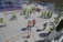 A group of people wearing safety vests and hard hats are gathered on a construction site. They are scattered around a flat concrete surface, with some engaged in conversation and others walking. In the background, there are scaffolding and construction materials, and several orange traffic cones are placed around the site.