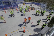 A group of people wearing safety vests and hard hats are gathered on a construction site. They are scattered around a flat concrete surface, with some engaged in conversation and others walking. In the background, there are scaffolding and construction materials, and several orange traffic cones are placed around the site.