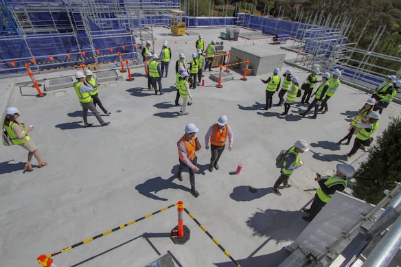 A group of people wearing safety vests and hard hats are gathered on a construction site. They are scattered around a flat concrete surface, with some engaged in conversation and others walking. In the background, there are scaffolding and construction materials, and several orange traffic cones are placed around the site.