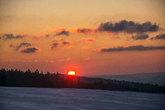 A stunning sunset over a forested landscape with a vivid orange sun descending behind a line of trees. The sky is painted in rich hues of orange, pink, and blue, with scattered dark clouds adding depth to the scene. The foreground features a snowy field, which contrasts with the warm colors of the sunset.