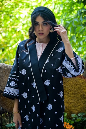 A person wearing a black robe adorned with white floral patterns stands in an outdoor setting. The robe has detailed embroidered designs on the sleeves and around the edges. The individual is wearing a turban-style headscarf, gold earrings, and a bracelet. Surrounding them is a lush green environment with blurred foliage in the background.