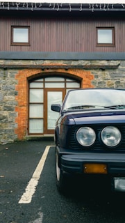 A classic car is parked on a black asphalt driveway in front of a building with a rustic stone and brick facade. The vehicle's headlights and part of the bumper are visible, and the building features a wooden door and two small windows on the upper floor.