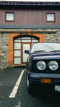 A classic car is parked on a black asphalt driveway in front of a building with a rustic stone and brick facade. The vehicle's headlights and part of the bumper are visible, and the building features a wooden door and two small windows on the upper floor.