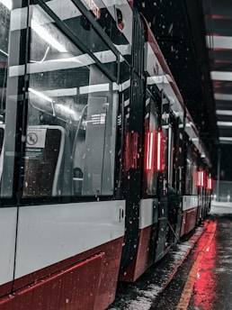 A modern tram or streetcar with red and white exterior is stationed indoors or at a covered platform. Snow is lightly falling, and the ground appears wet and reflective from melting snow. Red lights illuminate the interior and exterior, creating a contrasting glow against the dark surroundings. The scene is urban and captured at night or during low light conditions.