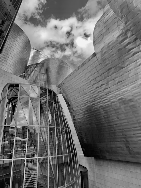 An architectural scene featuring a modern building with curved, metallic surfaces and large glass panels. The structure reflects an industrial and futuristic design, accentuated by the natural light and cloud-filled sky above.