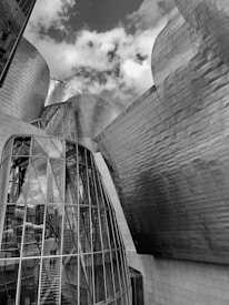 An architectural scene featuring a modern building with curved, metallic surfaces and large glass panels. The structure reflects an industrial and futuristic design, accentuated by the natural light and cloud-filled sky above.