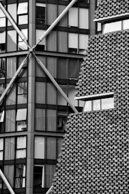 A detailed view of a building showcasing a contrast between two architectural styles. On the left, a modern glass facade is supported by visible metal beams arranged in a criss-cross pattern. On the right, a brick facade features a textured, staggered design, interrupted by a horizontal strip of windows.