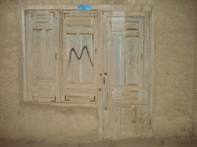 A rustic, weathered wooden door and window with faded paint, set into a rough plaster wall. The window has closed wooden shutters and the door features a simple panel design. There's a small, blue rectangular sign at the top, and a letter 'M' spray-painted in black on the shutters.