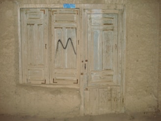 A rustic, weathered wooden door and window with faded paint, set into a rough plaster wall. The window has closed wooden shutters and the door features a simple panel design. There's a small, blue rectangular sign at the top, and a letter 'M' spray-painted in black on the shutters.
