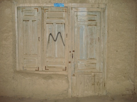 A rustic, weathered wooden door and window with faded paint, set into a rough plaster wall. The window has closed wooden shutters and the door features a simple panel design. There's a small, blue rectangular sign at the top, and a letter 'M' spray-painted in black on the shutters.
