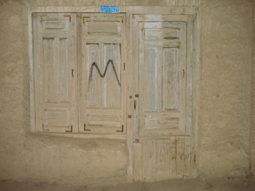 A rustic, weathered wooden door and window with faded paint, set into a rough plaster wall. The window has closed wooden shutters and the door features a simple panel design. There's a small, blue rectangular sign at the top, and a letter 'M' spray-painted in black on the shutters.