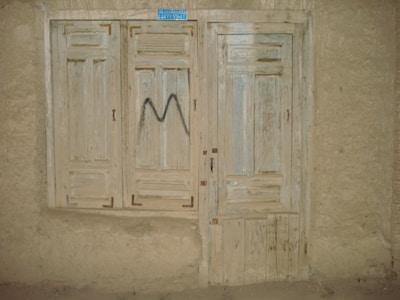 A rustic, weathered wooden door and window with faded paint, set into a rough plaster wall. The window has closed wooden shutters and the door features a simple panel design. There's a small, blue rectangular sign at the top, and a letter 'M' spray-painted in black on the shutters.